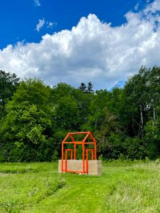 Large-scale outdoor sculpture consisting of a rammed-earth wall intersected by a bright orange steel framework in the outline of a house, installed within a grassy landscape surrounded by trees.