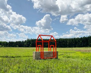 Large-scale outdoor sculpture consisting of a rammed-earth wall intersected by a bright orange steel framework in the outline of a house, installed within a grassy landscape surrounded by trees.
