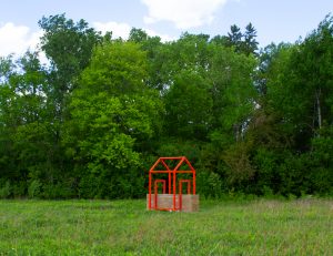 Large-scale outdoor sculpture consisting of a rammed-earth wall intersected by a bright orange steel framework in the outline of a house, installed within a grassy landscape surrounded by trees.