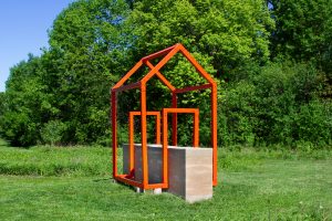 Large-scale outdoor sculpture consisting of a rammed-earth wall intersected by a bright orange steel framework in the outline of a house, installed within a grassy landscape surrounded by trees.
