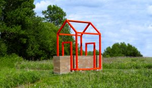 Outdoor installation view of How Much Earth For Sale?, a rammed-earth sculpture intersected by a bright orange steel house framework in an open landscape.