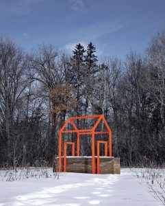 Large-scale outdoor sculpture consisting of a rammed-earth wall intersected by a bright orange steel framework in the outline of a house, installed within a grassy landscape surrounded by trees.