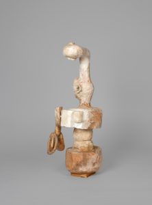 Hand-carved wooden sculpture composed of a vertical anthropomorphic figure with two carved heads connected by an elongated neck above a central torso. Projecting carved elements extend from either side of the body, and the surface is finished with stain, varnish, and wax that reveal the wood grain and tool marks.