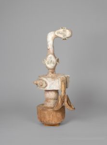 Hand-carved wooden sculpture composed of a vertical anthropomorphic figure with two carved heads connected by an elongated neck above a central torso. Projecting carved elements extend from either side of the body, and the surface is finished with stain, varnish, and wax that reveal the wood grain and tool marks.