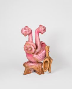 Hand-carved wooden sculpture composed of three interconnected anthropomorphic figures with elongated necks and simplified heads. A circular carved element and projecting wooden form extend from the side of the composition. Pink stain, ink, varnish, and wax emphasize carved textures while allowing the wood grain to remain visible.