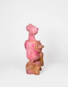 Hand-carved wooden sculpture composed of three interconnected anthropomorphic figures with elongated necks and simplified heads. A circular carved element and projecting wooden form extend from the side of the composition. Pink stain, ink, varnish, and wax emphasize carved textures while allowing the wood grain to remain visible.