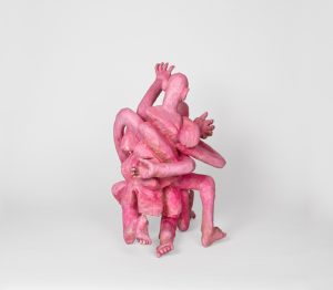 Hand-carved wooden sculpture composed of interconnected anthropomorphic figures with intertwined limbs and multiple heads. The compact composition is finished with stain, ink, varnish, and wax, revealing the wood grain beneath carved surfaces and visible tool marks.