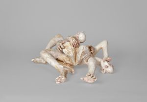 Hand-carved wooden sculpture composed of intertwined reclining anthropomorphic figures with elongated limbs, multiple heads, and overlapping bodies. The surface is finished with stain, ink, varnish, and wax, revealing carved tool marks and the natural wood grain.