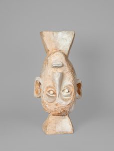 Hand-carved wooden sculpture of a bust with an elongated face, prominent ears, large circular eyes, and a tapered geometric form rising above the head. The surface is finished with stain, varnish, and wax, revealing carved facets, tool marks, and the natural wood grain.