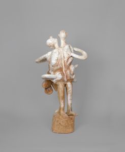 Hand-carved wooden sculpture composed of stacked anthropomorphic forms rising in a vertical arrangement with multiple heads and projecting limbs. Metal elements integrate with the carved wood, while stain, ink, varnish, and wax emphasize the wood grain and hand-carved surface