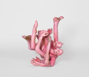 Hand-carved wooden sculpture composed of intertwined anthropomorphic forms with multiple heads, arms, and legs arranged in a compact, inverted-looking configuration. Pink stain, ink, varnish, and wax emphasize the carved surface, wood grain, and visible tool marks.